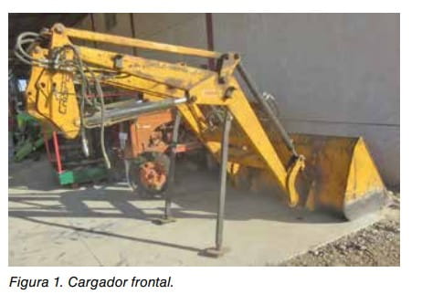 Figura 1: cargador frontal agrícola con brazo articulado y cazo, apoyado junto a una nave.