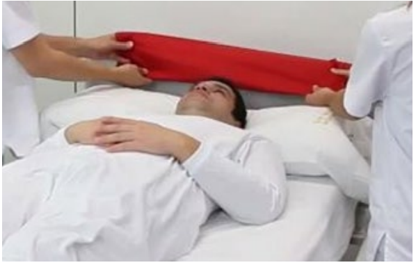 Dos personas deslizan una sábana roja bajo la espalda de un paciente tumbado en una cama para su movilización
