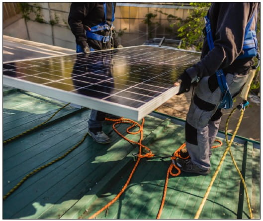Dos operarios con arnés trasladan un panel solar sobre una cubierta inclinada con líneas de vida
